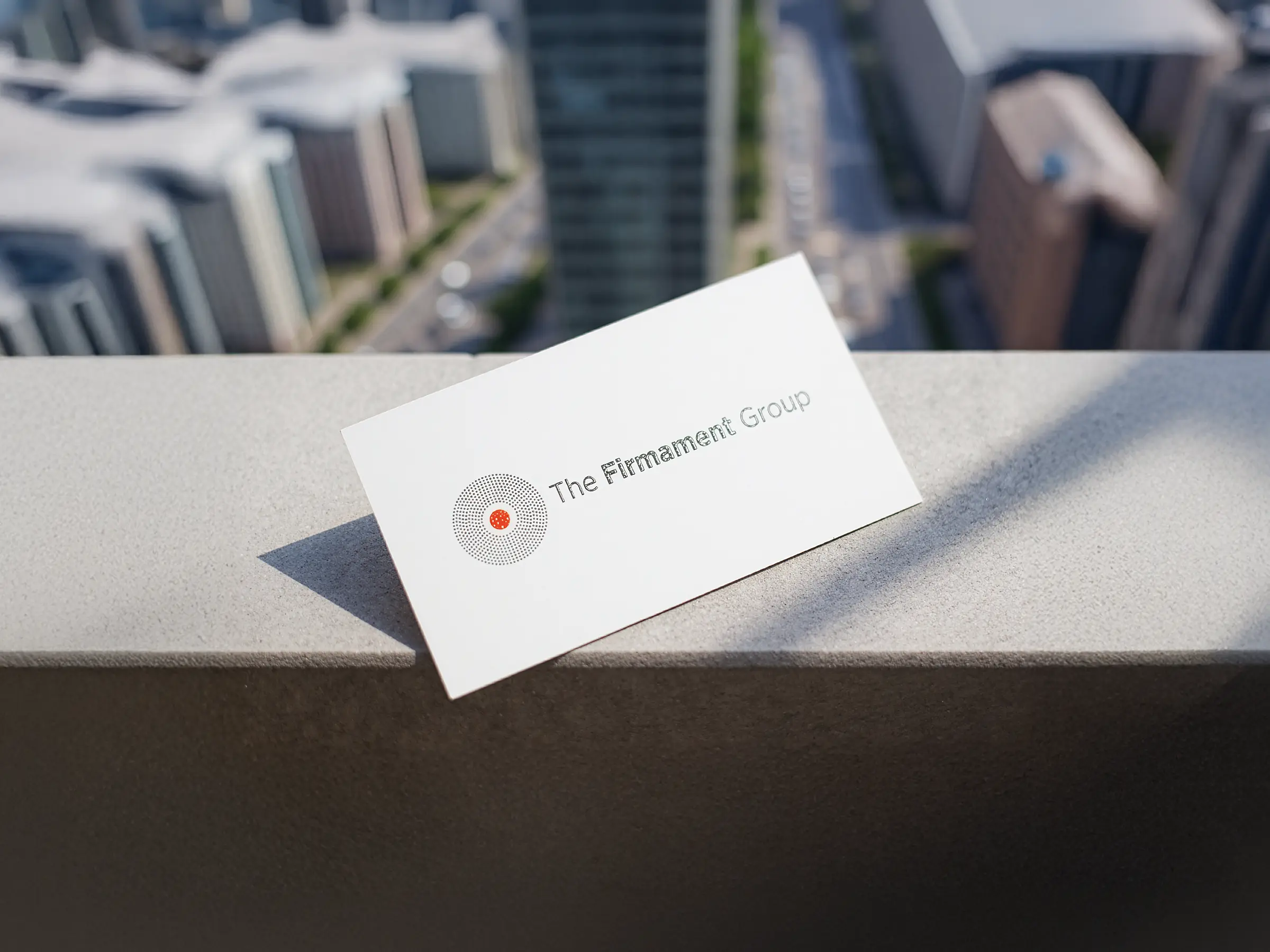 A premium white business card for The Firmament Group featuring a textured circular logo and raised ink, custom-printed at our NYC shop and displayed on a stone ledge overlooking the city.