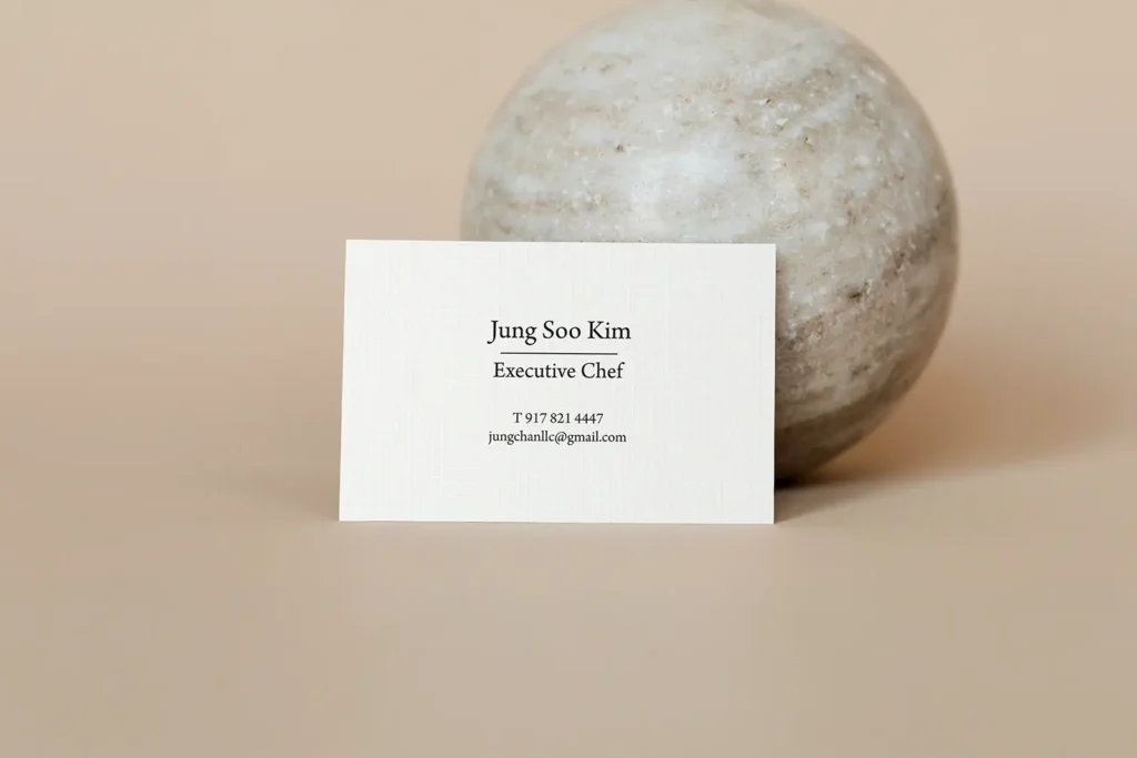 Minimalist executive chef business cards on premium white linen cardstock with a subtle texture, propped against a stone sphere to emphasize elegant, simple design.